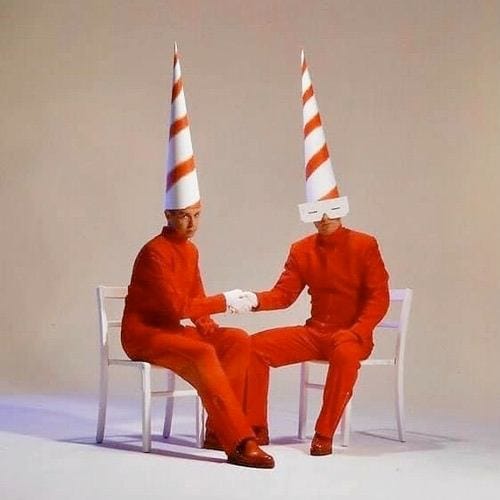A picure of the synth pop due, The Pet Shop Boys in 1993, pictured in odd garb of red jumpsuits, with conical dunce type hats, sitting and shaking hands oddly.