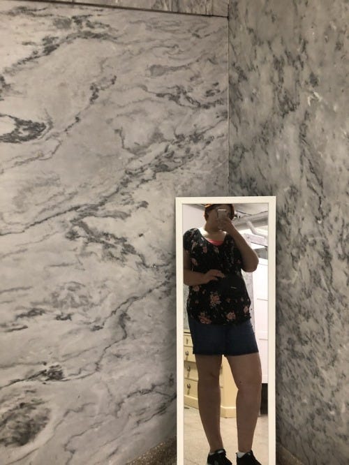 A white woman in her mid-twenties is standing in front of a mirror, which has been placed against a marble-patterned wall. She is holding the phone such that her face is obscured. She is wearing a black floral-patterned blouse and dark jean shorts with sneakers. There is a small black purse on her shoulder.