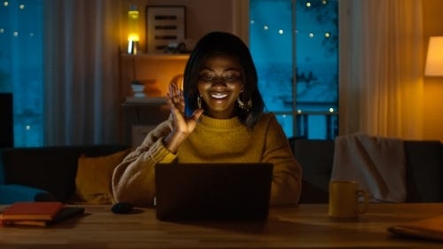 A person in a yellow jumper sits in front of a laptop which illuminates their face. They have their right arm raised towards their face, as if while talking to someone on the screen. In the background is a couch, window, and shelf.