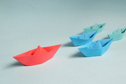 One red paper boat followed by two blue and two green paper boats