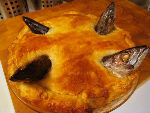 Image of a pie with pilchard heads poking out from the top crust. The pie is in a round dish on a wooden table