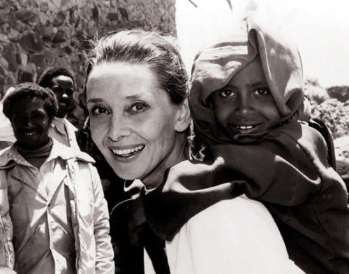 Audrey Hepburn in Africa. She is looking at the camera and is wearing a white shirt. An African child is riding on her back.