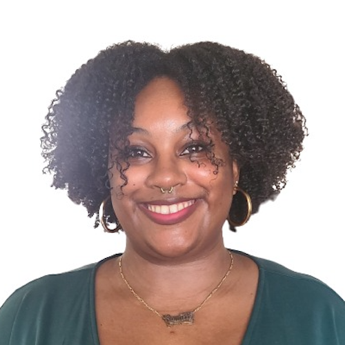 Headshot of Camille Williams, a Black brown-skinned femme, from the shoulders up. She has on pink lipstick and is looking at the camera with a wide smile. She has black curly hair, winged liner, a gold septum ring, gold hoops, and a green top with a gold name-plate necklace.