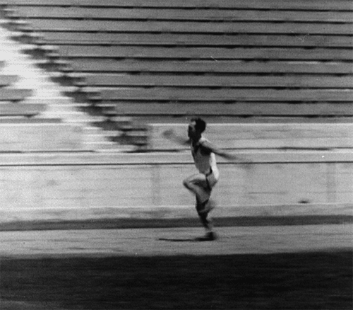 #praCegoVer homem em um filme preto e branco correndo em uma pista de corrida com obstáculos, ele tropeça, mas continua