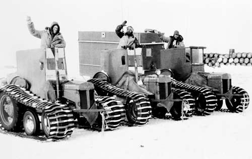 Black and white photograph of the Ferguson tractors in Antarctica in the snow. They are lined us together. A driver is waving from the top of the cabin on each tractor.