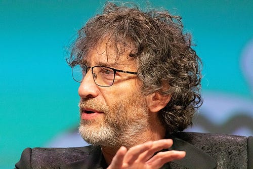 Image of author Neil Gaiman, an older white man with curly graying brown hair, a short beard, and glasses. He appears from the shoulders up wearing a suit, gesturing as if in the middle of explaining something, against a blue background.