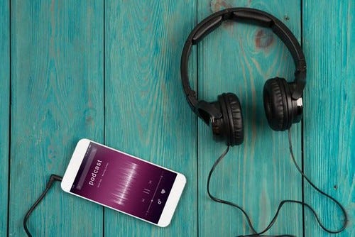 A photo of a pair of headphones plugged into a smartphone, on a wooden table. The smartphone is playing a podcast.