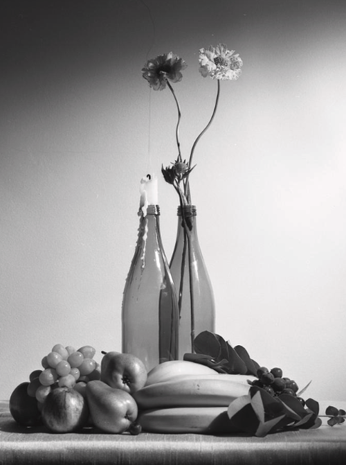Still Photography of two bottles, one with two flowers and the other with a candle. The bottles are surrounded by fruits.