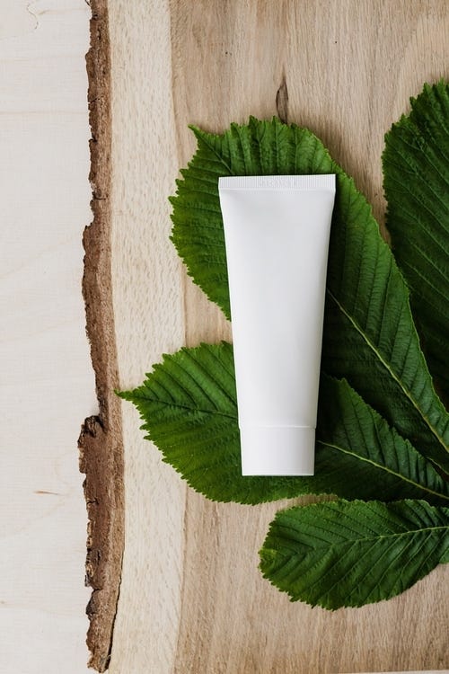 A blank white product tube lying atop green leaves and grainy wood.