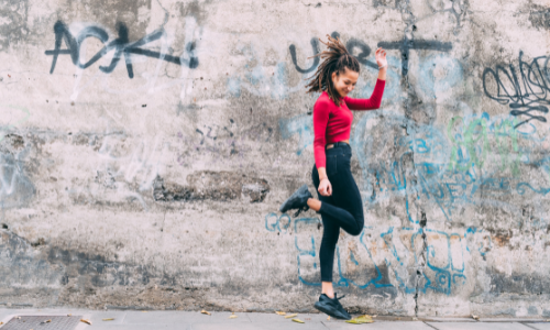 young woman in red shirt leaping in joy in front of a wall with graffiti