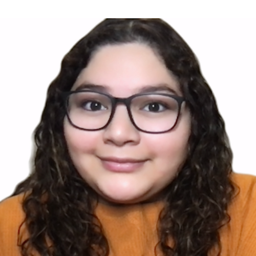 A photo of a Latina woman, Ruby, from the shoulders up. She is facing the camera with her head slightly tilted, and a closed-lipped smile upon her face. She is wearing an orange long sleeve top, no jewelry in sight, with black glasses. Her cheeks have a hue of pink and her dark brown hair is in its natural, curly form.