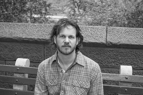 black-and-white photo of Adam Giannelli, a white man with a short beard and medium-length hair. He wears a plaid shirt and looks calmly at the camera.