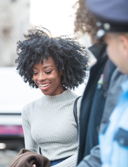 Una mujer negra con un suéter de cuello alto color crema mira hacia abajo y sonríe ampliamente mientras un oficial de policía