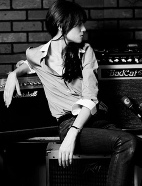 A black and white photograph of a tomboyish woman. She is sat in profile against some speakers.