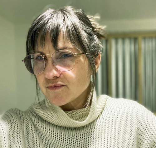 Jennifer Parker is a white woman wearing a beige turtle-neck shirt and light brown rim glasses. Her brown hair is in a bun and she has bangs resting over her forehead. In the background looks like a long window with light brown frames and a light blue curtain.