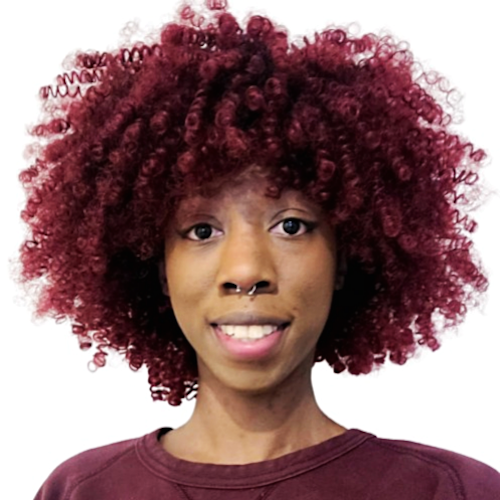 Photo of Pandora, a Black nonbinary femme, from shoulders up. They are looking at the camera with an open, lipped smile. They are wearing a purple sweater and their hair is red.