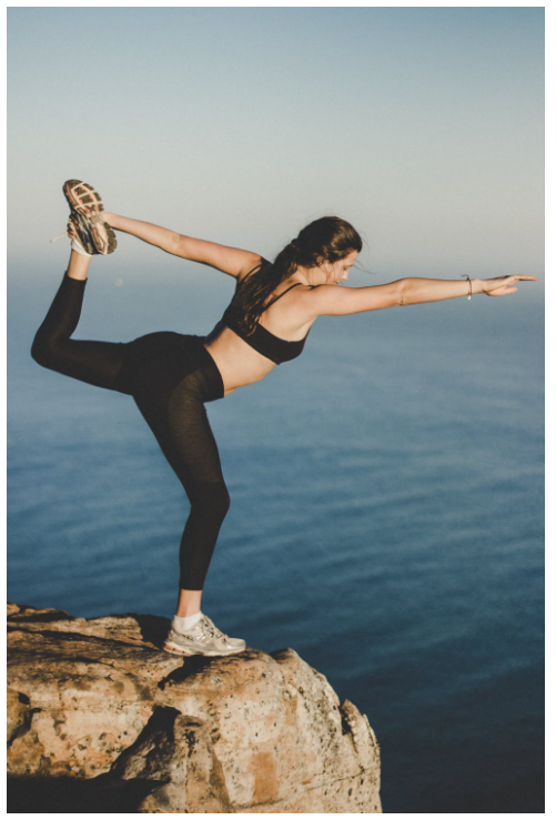 A woman is on a cliff, dressed in black tights and sports bra, doing stretched out yoga pose, overlooking a body of water.