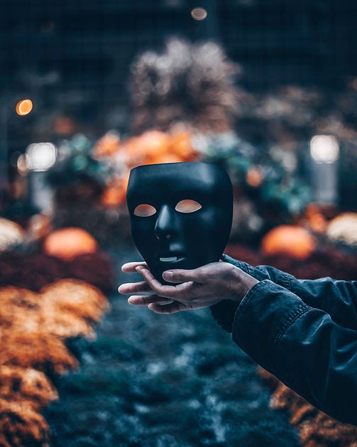 A pair of hands holds a black mask, the symbol of who society believes they are. The actually person stands out of frame. Afraid to expose themselves to the world without their mask.