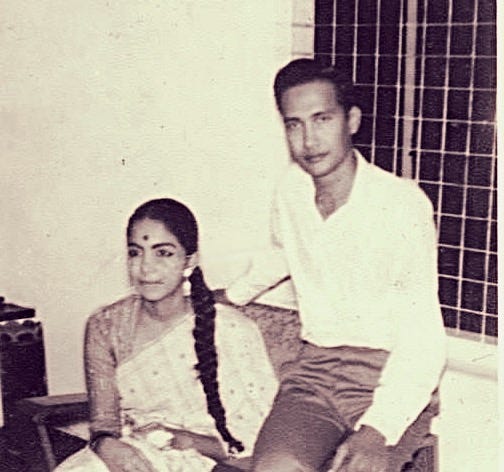 A faded, black-and-white picture shows an Asian woman in a sitting in a chair. She has a long ponytail. Seated on the arm of a chair is a lanky man. He is clean-shaven, wearing a white shirt and black pants and smiling at the camera.