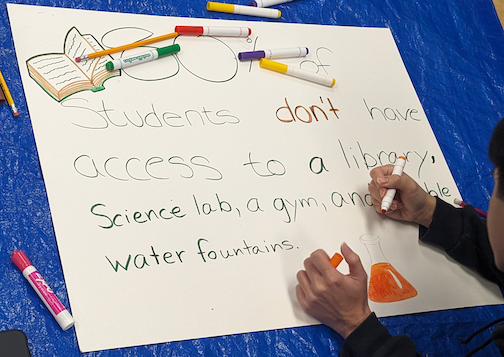 Photo of a student making a sign: “Students don’t have access to a library, a science lab, a gym, and useable water fountains.”