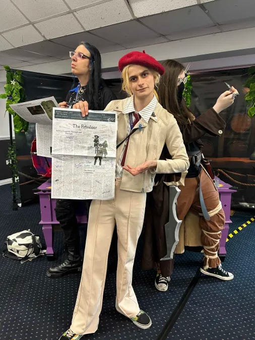 Three individuals stand posing together, each attempting to mimic their pirates’ poses. In the middle is Belle, is holding up a broadsheet sized newspaper titled ‘The Privateer’. She has bleach blonde hair and wears a beige outfit that is compromised of trousers and a jacket and a shirt. On the right is Jess, who is in a cosplay of her pirate. She has glasses and a face covering, and brown hair. To the left is Dray who is wearing mostly black, save for a few blue accents. They wear glasses.