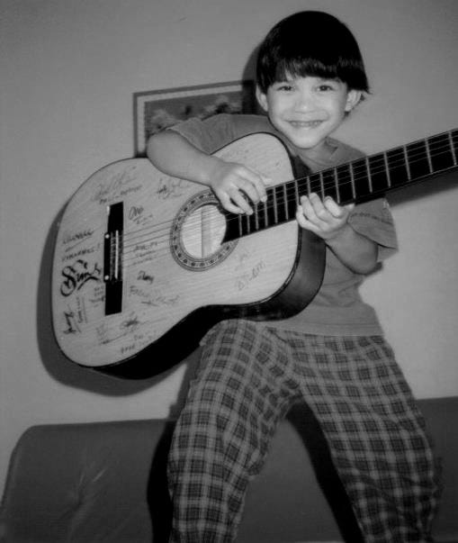 Emillio as a child, smiling while holding a guitar, standing on a bed in his pyjamas.