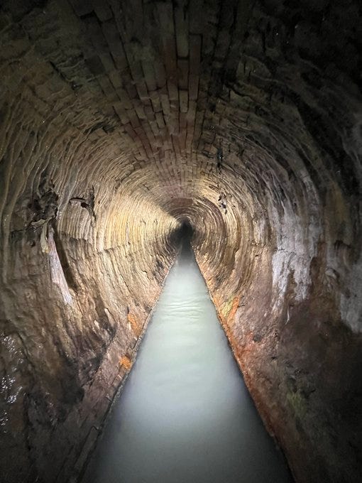 Long stretch of sewer under Cleveland. Brick lined and built in 1873 it is still in use today. Brick is rounded and very weathered but intact. walls are lined with white mineral deposits and a stream of discolored water is flowing down the bottom middle of the pipe. photo credit Matt Barker, Cody Kelley, and Damien Csiszar.