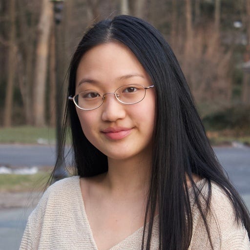 Headshot of a young woman with long dark hair and glasses gently smiling at the camera. The background has out-of-focus nature scene