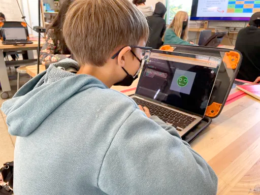 Photograph of a student working at a laptop. On the screen is an illustration of a green face with eyes and a mouth, to the left of the screen is the coding window that generated the image.