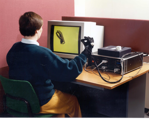 Someone sitting in front of a computer wearing the dataglove, which is mirrored with a 3D model on the screen.