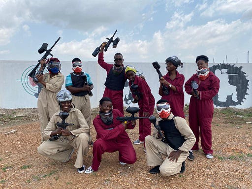 Nine people in red and beige overalls, googles and vests pose while holding paintball guns.