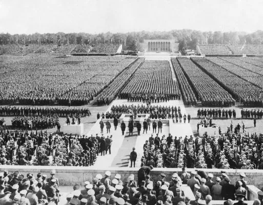 A imagem mostra uma grande multidão reunida em um enorme comício nazista. É um ambiente ao ar livre e a multidão parece estar participando. A foto está em preto e branco.