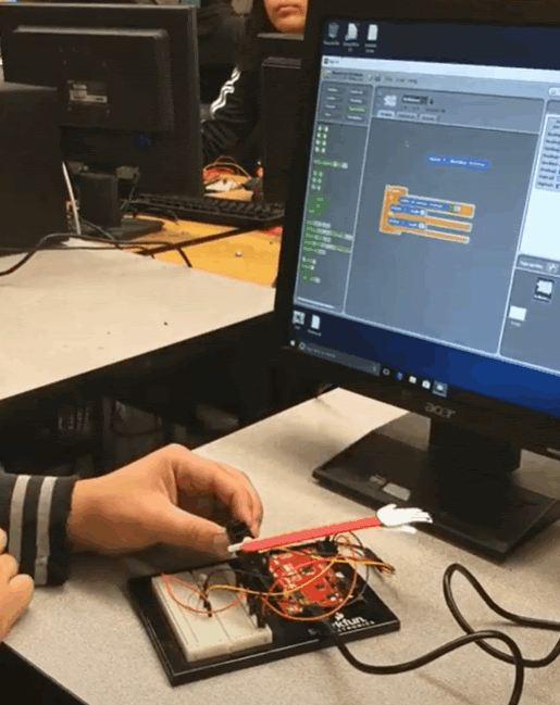 Arduino project in front of a computer. A person puts their hand to it and a popsicle stick w/ a small paper hand high-fives
