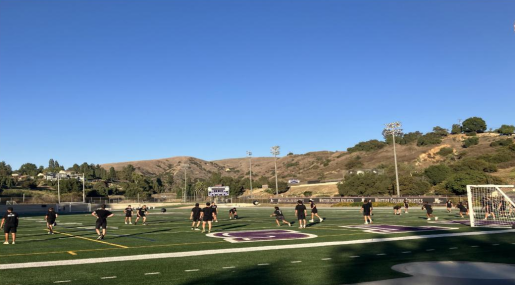 During a day with a clear, blue sky, many women are on a football field. They appear to be in the middle of practice and are wearing black uniforms and white shoes. They are in hills and are on the side of the football field with a big purple letters.