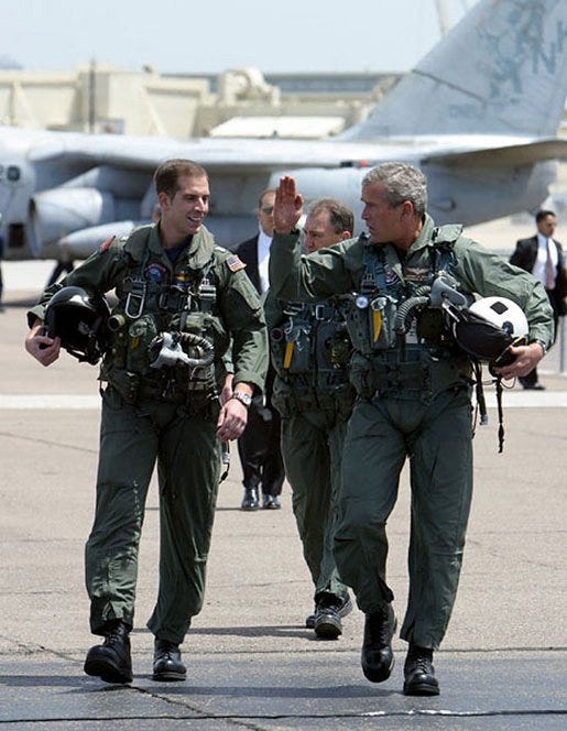Bush talks with a pilot while both are in pilot uniforms and walking on tarmac.