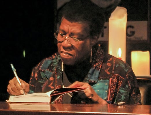 Octavia Butler seated and signing one of her books. She’s wearing glasses and a patterned shirt, and has a short well-maintained afro hairstyle. There are candles and part of a sign visible in the background.