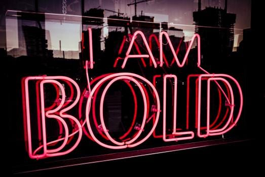 A red neon sign in a window that reads “I AM BOLD” with tall buildings reflected in the background