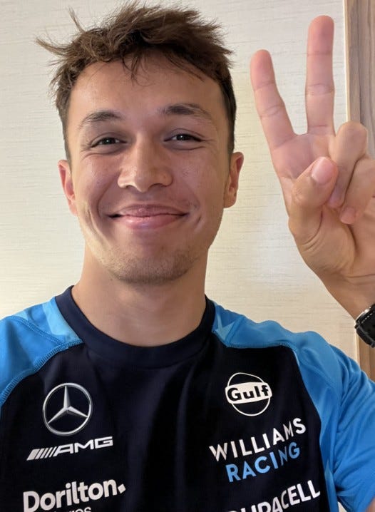 Alex Albon grinning and giving the ‘peace’ sign wearing his Williams team shirt.