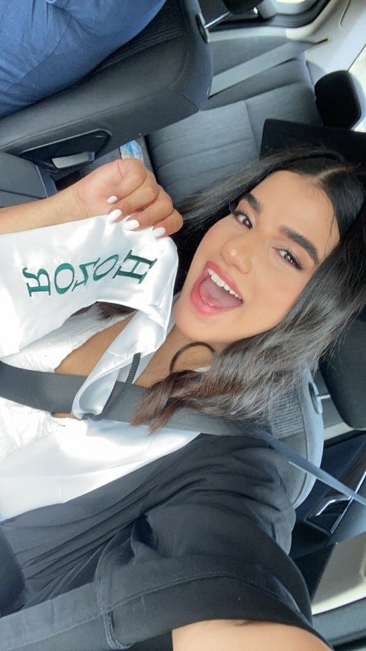 Sarah taking a selfie and smiling while holding up a white sash that says “honor.”
