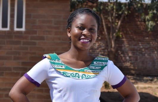 A young Malawi woman smiles with her hands on her hips. She wears a white shirt with a collar