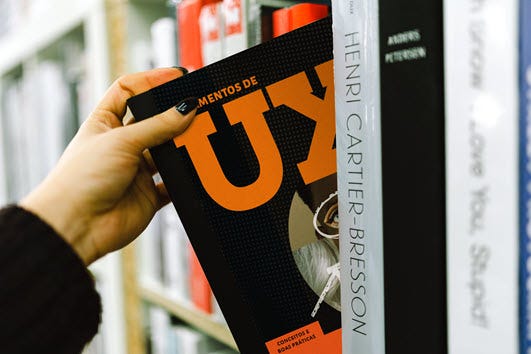Foto de uma estante de livros com uma mão retirando o lvro Fundamentos de UX da prateleira.