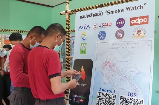 Two men in matching outfits look at their smartphones while standing in front of a large sign titled “Smoke Watch” and including several agency logos, a large map, and QR codes for Android and IOS devices.