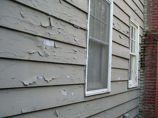 Gray wood siding with peeling paint and two white windows.