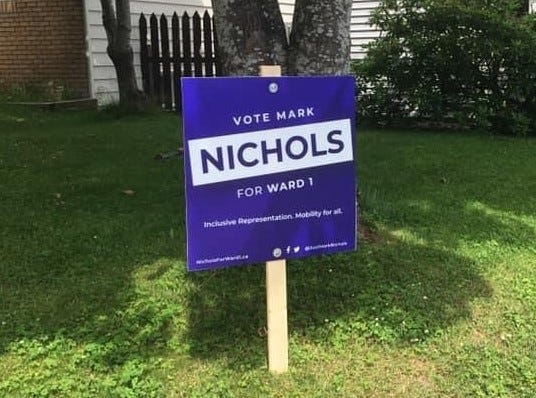 A purple campaign sign on someone’s lawn that says, “VOTE MARK NICHOLS FOR WARD 1”. IN smaller print it says, “Inclusive Representation. Mobility for all.”