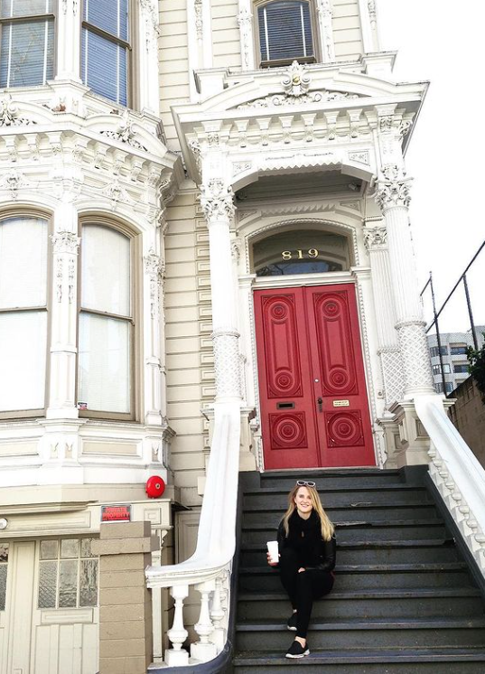 A woman holding a coffee sits on the steps of a building with a red door.