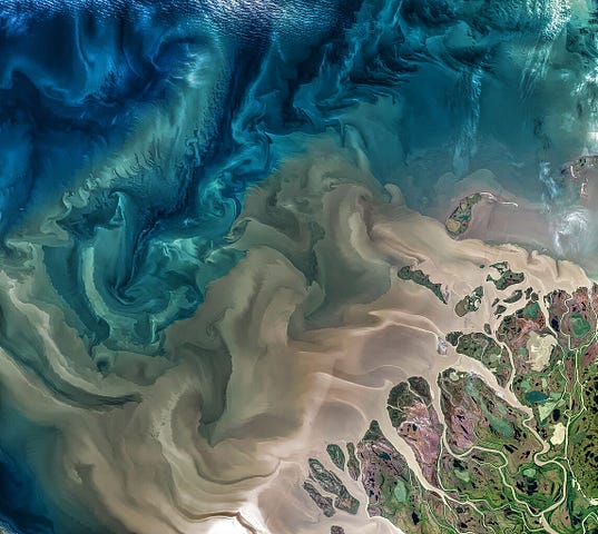 A NASA full colour satellite photo of a river delta, with swirling blues and yellows and branching tributaries on land in the bottom right corner of the image