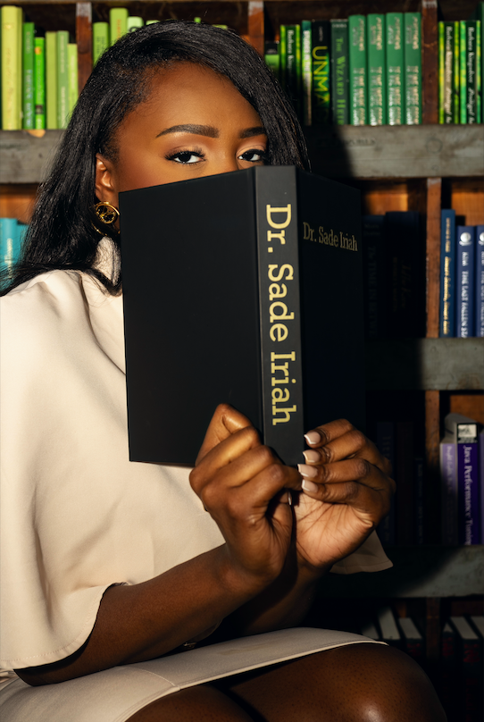 A Black woman holding a black book that has &lsquo;Dr. Sade Iriah&rsquo; in gold letters on the spine of the book. She is covering the bottom half of her face with the book and you can see the top portion of her face. She has a cream dress that has drape-like sleeves. She in front of a bookshelf in a library. She has french tip manicured nails.