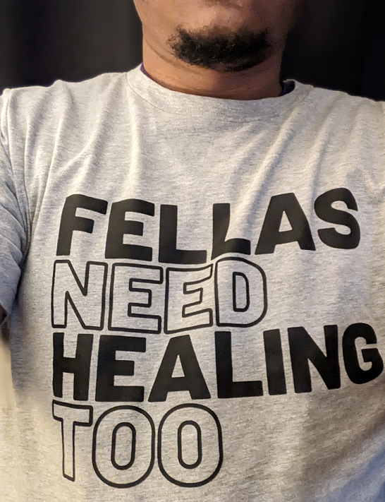 Black man wearing shirt that reads “Fellas Need Healing Too”
