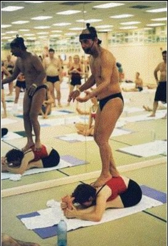 Subject seated on floor in yoga class, folded forward; big toe touching forehead. Male instructor standing on her back.