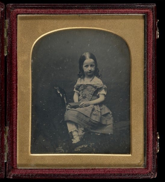 Black and white daguerreotype photo of a little girl in an elaborate dress. The photo is in a metal frame in a leather case.